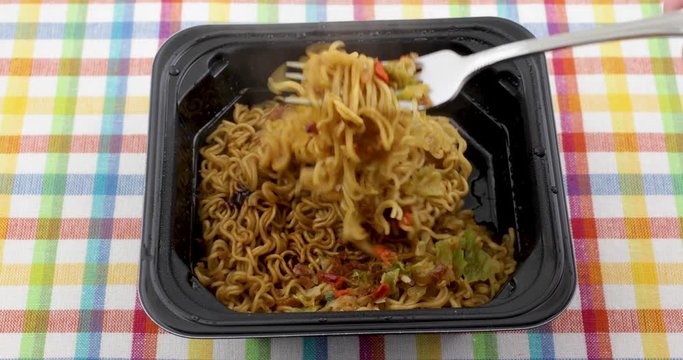 Video of mixing a microwaved beef flavored ramen noodles meal with a fork in a black tray atop a colorful place mat.