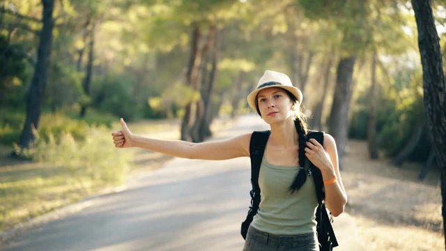 Traveler woman hitchhiking on a sunny forest road. Tourist girl looking for ride to start her journey
