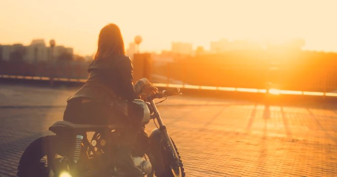 Beautiful Caucasian female biker sits on a custom built cafe racer motorcycle on a rooftop parking lot, enjoying beautiful sunset over city. 4K UHD 60 FPS