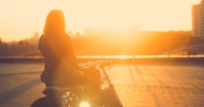 Beautiful Caucasian female biker sits on a custom built cafe racer motorcycle on a rooftop parking lot, enjoying beautiful sunset over city. 4K UHD 60 FPS