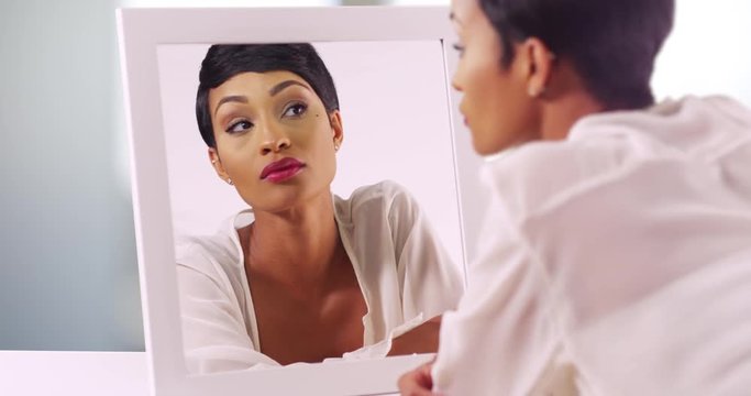Gorgeous black female in her 20s gazing at reflection in the mirror. Close up of African American woman with pixie haircut looking at reflection inside bathroom 