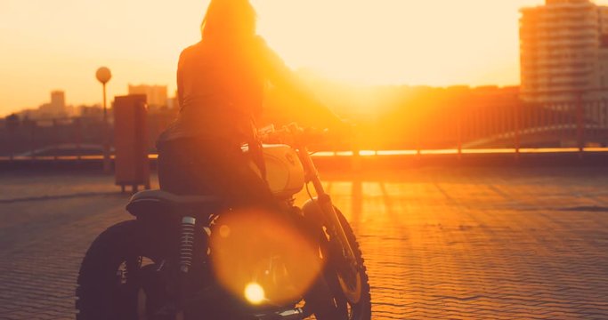 Beautiful Caucasian female biker dismounts her custom built cafe racer motorcycle on a rooftop parking lot, beautiful sunset over city in the background. 4K UHD 60 FPS