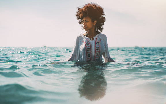 Attractive young black curly girl in traditional African chemise is standing up to the waist in ocean water and looking aside, strong reflection in the bottom, ripples on water surface