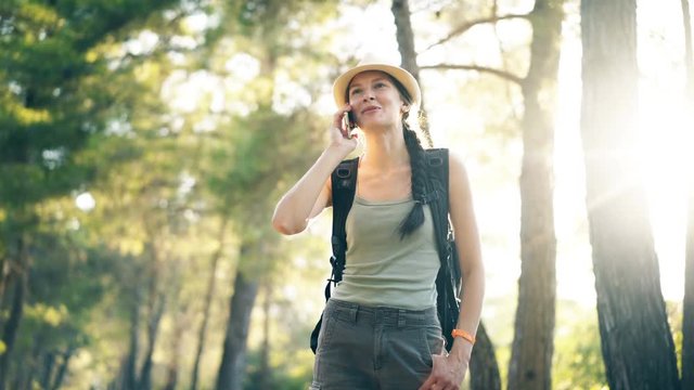 Young tourist girl backpacker in hat chatting with smartphone during walking in summer forest