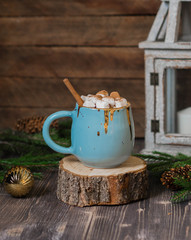 Large blue ceramic mug with hot chocolate, marshmallow and cinnamon stick on a New Year's background with a candlestick, spruce branches and a Christmas tree toy
