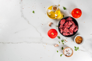 Meat, beef. Fresh raw chopped goulash, beef cubes in a bowl. Spices (salt, pepper), tomatoes, garlic, onions. On a white marble table, with a fork for meat and a knife. Top view copy space