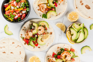 Seafood. Mexican food. Tortilla tacos with traditional homemade salsa salad, parsley, fresh lemon, avocado and grilled shrimp pawns. On a white marble background. Top view copy space