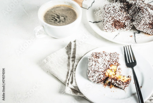 Australian food. Traditional dessert Lamington - pieces of biscuit in dark chocolate, sprinkled with coconut powder chips. On a marble plate, white table. With coffee mug. Copy space
