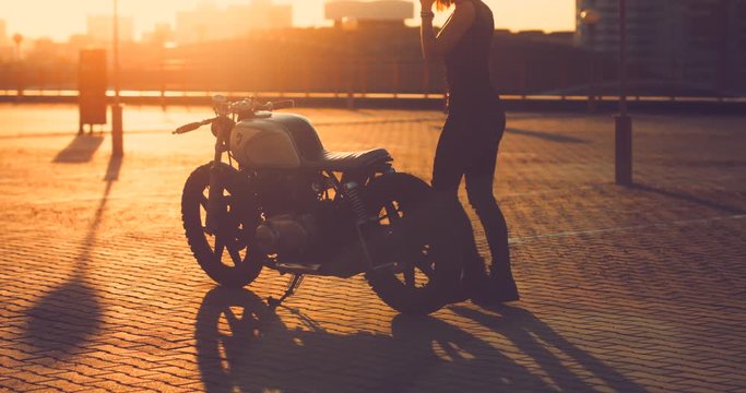 Beautiful Caucasian female biker mounts her custom built cafe racer motorcycle on a rooftop parking lot, beautiful sunset over city in the background. 4K UHD 60 FPS