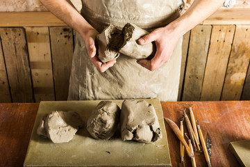 pottery, clay, ceramics art concept - top view on hands of young master holding the large pieces of fireclay, male dressed in an apron, a ceramist with raw materials on table with sculpting tools set