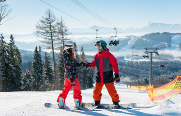 Pair of snowboarders on top of a ski slope at winter resort downhill on a sunny day. Man holds a woman's hand against the backdrop of a ski-lift and snow-capped mountains. Bukovel © anatoliy_gleb