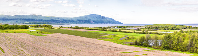 Fototapeta na wymiar Panorama, panoramic aerial view of farmland in Ile D'Orleans, Quebec, Canada, plowed field, furrows, land, farm, house, barn, shed, Saint Lawrence river, hills, mountains and distant village