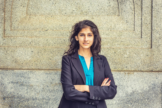 Modern East Indian American Student. Dressing in black blazer, blue under shirt, crossing arms,  a young girl with long curly hair standing by wall, smiling, looking at you. Instagram filtered effect.