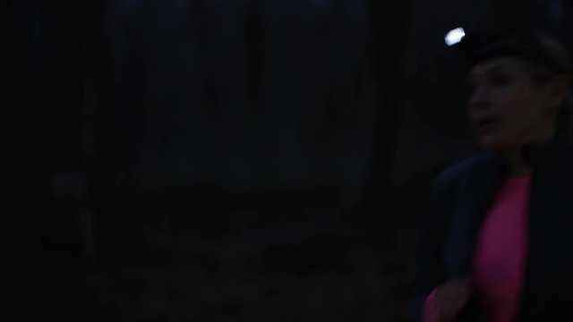 Woman on night run in the woods, wearing lamp on headband, stops to check smartwatch.