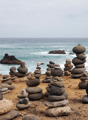 Fototapeta na wymiar piles of stacked stones and pebbles in zen towers on a yellow sandy beach with sea breaking over rocks and blue calm sky
