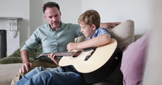 Father teaching son guitar chords at home on sofa