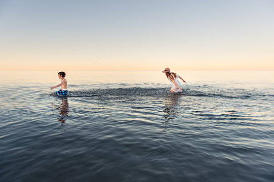 Girl chasing and splashing her brother in calm water at dusk