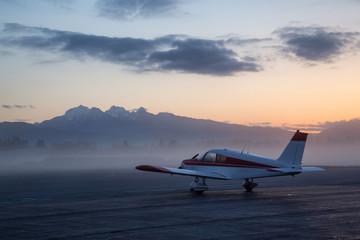 Fototapeta na wymiar Airplane parked at the airport during a colorful and vibrant sunrise with mouhtains in the background. Taken in Pitt Meadows, Greater Vancouver, BC, Canada.