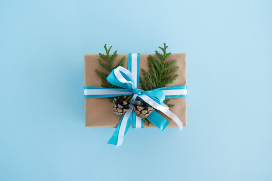 Gift box wrapped of craft paper, blue and white ribbons and decorated fir branches and pinecones on the blue background, top view. Christmas present.