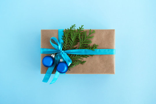 Gift box wrapped of craft paper, blue ribbon and decorated fir branches and blue Christmas balls on the blue background, top view. Christmas present.