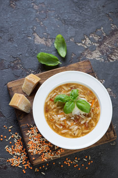 Italian soup with lentils, pasta and parmesan cheese on a wooden serving board. High angle view on a brown stone background with space