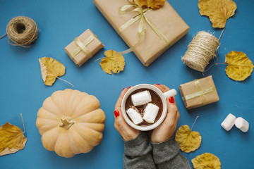 Woman hands with hot cocoa with marshmallows, gifts, packing paper, dry leaves on a blue table. top view, flat lay, autumn concept