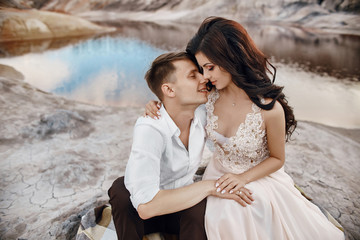 Fototapeta na wymiar Beautiful couple in love kissing and hugging sitting on the rocks amid the fabulous mountains. Red blood lake. A man and a woman in a beautiful wedding dress. Long light summer dress on a woman body