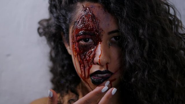 Scary portrait of young girl with Halloween makeup smears blood on her face. Beautiful latin woman with curly hair looking into camera in studio. Slow motion.