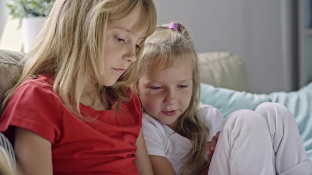 Tilt down of cute little girl with blond hair and her little sister sitting on sofa and chatting while playing on tablet