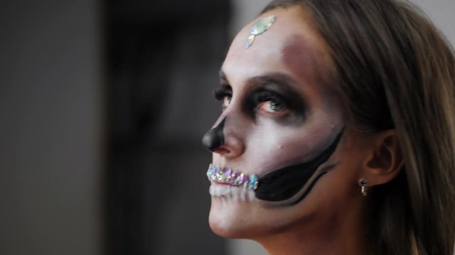 Scary young girl with creative halloween face art in the dressing room. Portrait of glamorous skull with rhinestones and sequins. Professional greasepaint for the celebration
