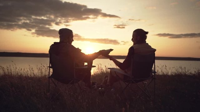 Two men friends camping near a river during sunset, passing food from a parcel. Slow motion.