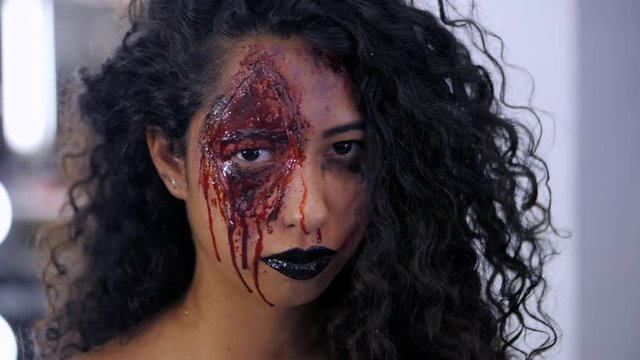 Scary portrait of young girl with Halloween blood makeup. Beautiful latin woman with curly hair looking into camera. Slow motion.