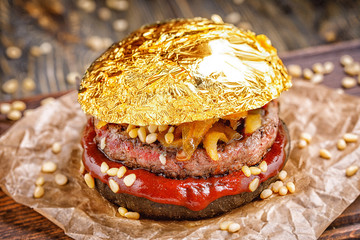 Gold beef burger with a black bun,with arugula and cheese and ketchup sprinkled with pine nuts served on pieces of brown paper on a rustic wooden table, on a dark background. Delicious gold burger