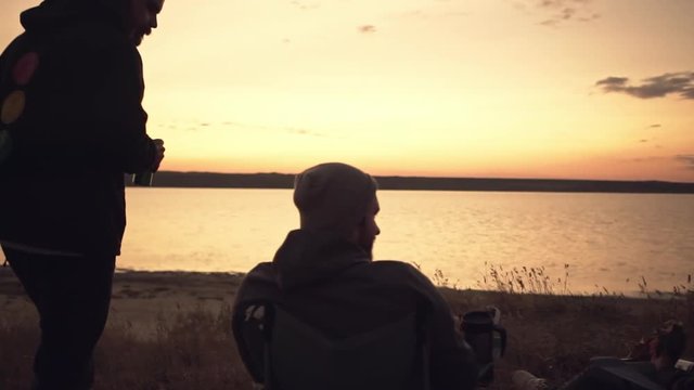 Group of friends watching sunset, camping together near water. Slow motion.