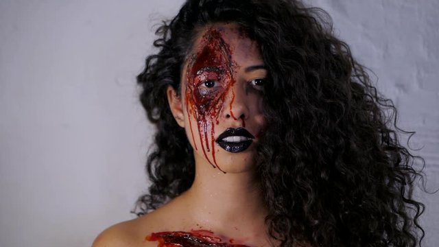 Scary portrait of young killer girl with Halloween blood makeup. Beautiful latin woman with curly hair looking into camera in studio. Slow motion.