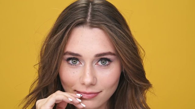Close up view of beddable shy brunette woman touching her face and looking at the camera over orange background