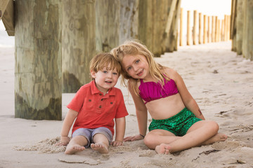 adorable brother and sister siblings sitting under dock pier on beach 