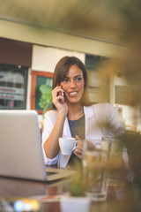 Young smiling business woman working in cafe talking on phone and holding cup of coffee.