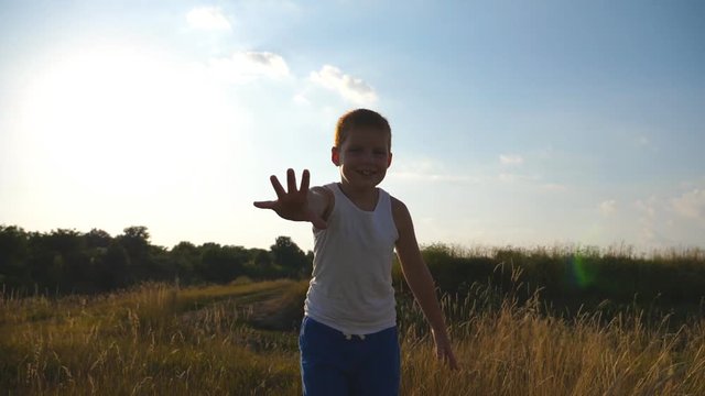 Happy little boy with an outstretched hand running at field behind camera and trying to catch it. Smiling male kid having fun in nature on a summer meadow. Child jogging outdoor. Slow motion
