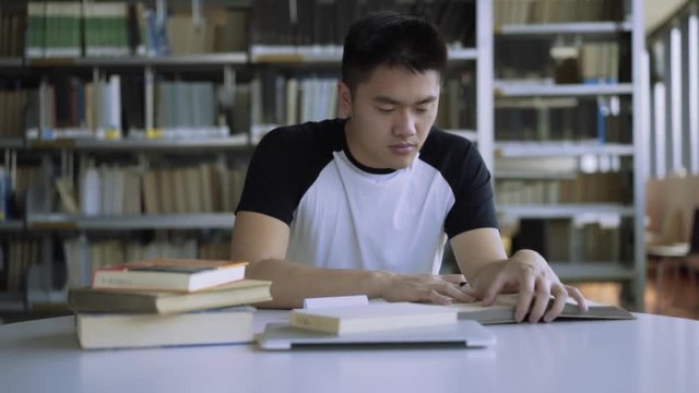 Tracking shot of student studying at desk in library in college. Focused student prepares for his exam in library. Book shelves in background