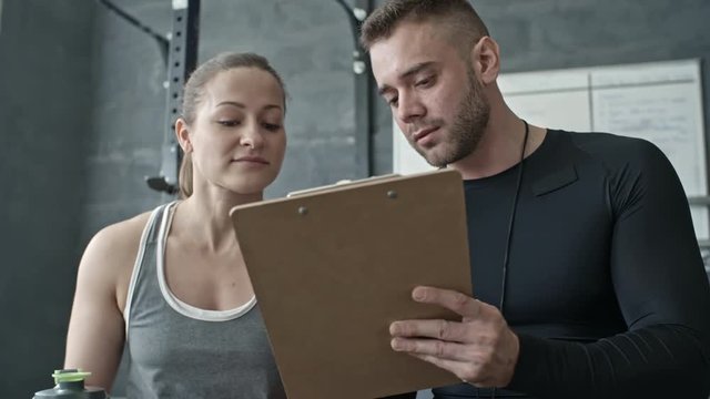 Muscular personal coach writing on clipboard and talking to young woman in cross training gym