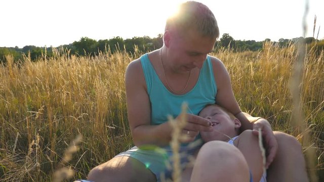 Happy young man sitting on grass at the field and playing with his baby boy. Cheerful daddy and his child having fun together in nature. Father tickling his little son outdoor. Slow motion Close up