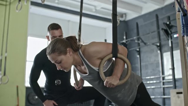 Slowmo of young woman doing ring push-ups in cross training gym with support of personal coach