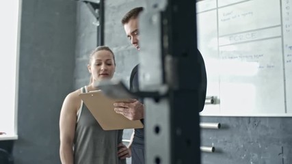 Young handsome personal coach holding clipboard and instructing sporty woman before cross training in gym