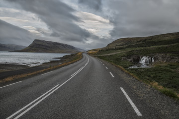 a deserted asphalt road running away into the hills, along the road a waterfall. Iceland. The spirit of travel and adventure.
