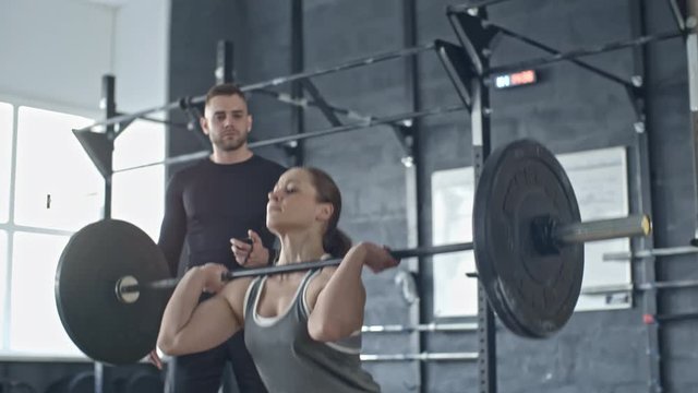 Young coach looking at muscular woman doing exercise with weighted barbell in cross training gym