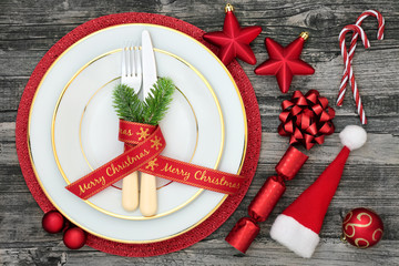 Christmas table place setting with dinner plates, napkin, cutlery, fir, ribbon, santa hat, candy canes, cracker and bauble decorations on rustic wood background. - Powered by Adobe