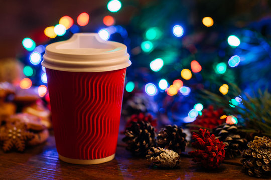Delicious Christmas with take away latte. Close up cup of warm energy drink and strobila decoration on festive fairy lights background. Cozy xmas evening in cafe and fairs, happy holiday concept