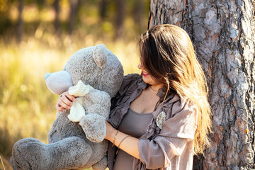 portrait of a beautiful young pregnant girl in a fashionable dress against the backdrop of an autumn forest, holding a teddy bear, a stuffed toy