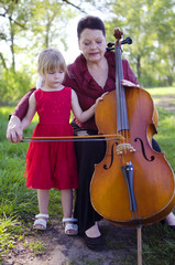 Old women and blond caucasian girl playing concert on cello outdoors. Happy grandmother teaching to play violoncello her granddaughter.